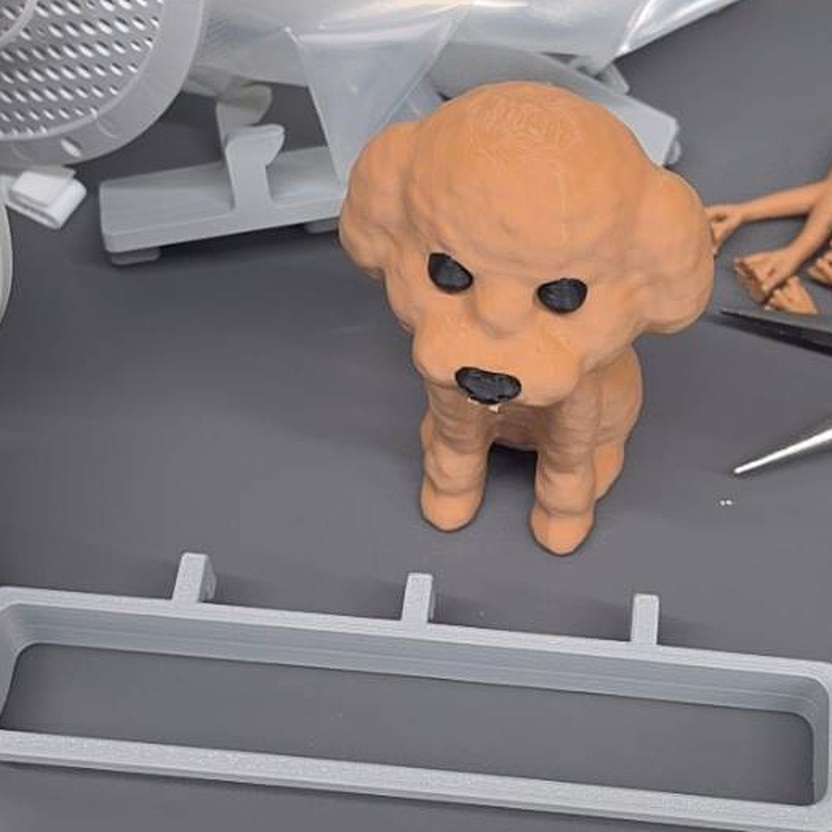 オレンジ色の樹脂で出力したトイプードルの置物と、奥の机に置かれた工具や樹脂の巻き糸が写る写真